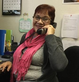 Virginia answers the phone at her office desk, wearing a bright pink scarf, glasses, a gray sweater and shirt, and has short, red hair.