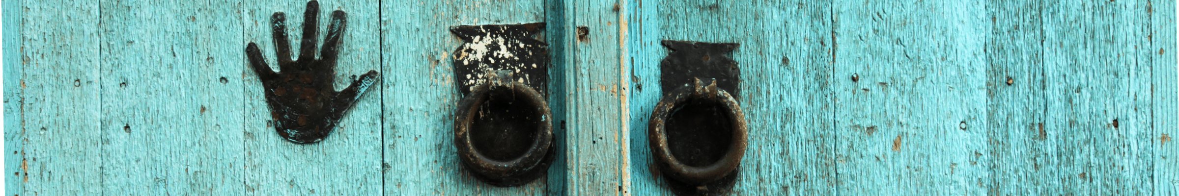 Turquoise painted wooden door with two iron door handles and an small outline of an iron hand placed on the left side. 