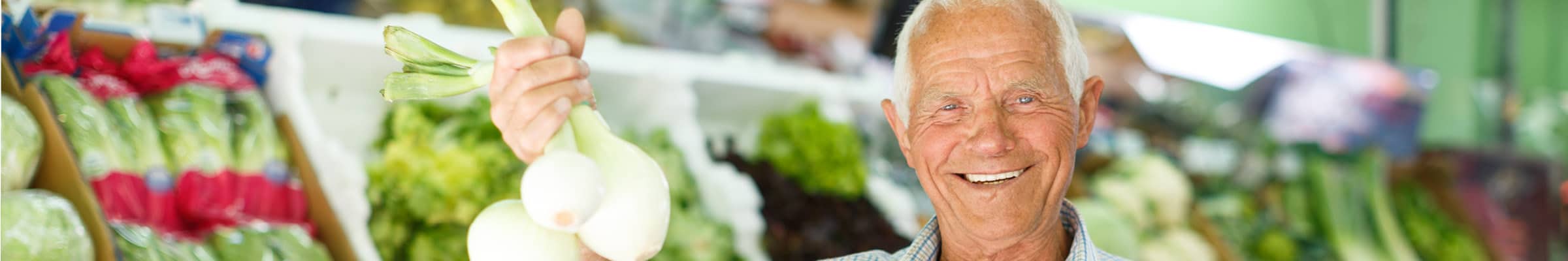 Older male is smiling and holding up a bundle of leeks in his hand, standing in the produce section of a grocery store.