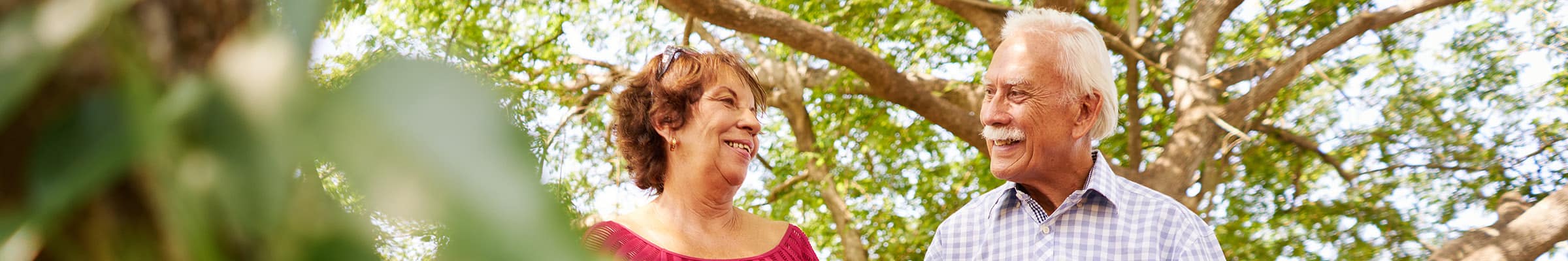 Older hispanic male and female smiling at each other while outside beneath green trees walking.